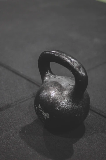 A sleek black kettlebell resting on a dark textured gym floor under moody lighting.