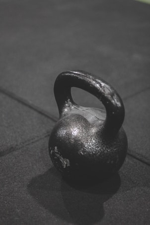 A heavy black kettlebell rests on a dark, textured surface, casting a shadow beneath it. The kettlebell has a slightly worn appearance with patches of paint chipped off, indicating frequent use.