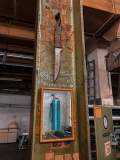 A vintage knife with a wrapped handle is mounted on an industrial-style metal surface, alongside a small framed mirror. The background shows a workshop environment with various industrial equipment and clutter, including pipes and machinery. The surface around the knife is adorned with faded and torn stickers.