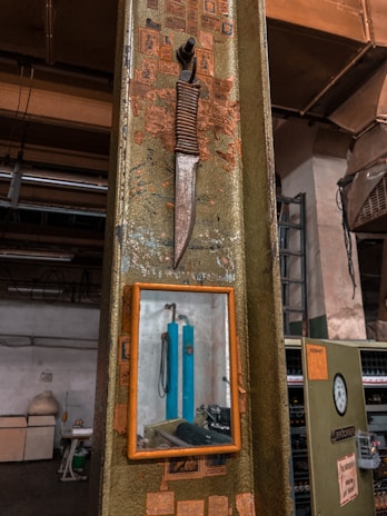 A vintage knife with a wrapped handle is mounted on an industrial-style metal surface, alongside a small framed mirror. The background shows a workshop environment with various industrial equipment and clutter, including pipes and machinery. The surface around the knife is adorned with faded and torn stickers.
