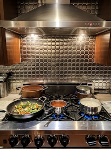 A kitchen stove features multiple pots and pans cooking various dishes. On the left, a frying pan contains sautéed vegetables including broccoli, carrots, and green beans. In the center, a saucepan is filled with a tomato-based sauce. To the right, a pot is boiling possibly pasta or rice. The stove is stainless steel with several burners lit, and there's a modern metallic backsplash with a grid pattern. Above, there's a large, silver range hood.