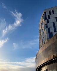 A modern building with glass and metal panels against a backdrop of a clear blue sky with wispy clouds. The building features large windows and a curved facade.