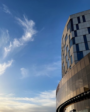 A modern building with glass and metal panels against a backdrop of a clear blue sky with wispy clouds. The building features large windows and a curved facade.