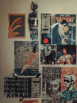 A collection of vintage posters is displayed on a wall, including a mix of music and artistic themes. One poster features a stylized graphic design with abstract shapes, while another depicts a person wearing a striped shirt. There are several concert posters, one of which advertises a performance by Dee Dee Ramone, showcasing a musician with a guitar. Another poster features an artistic depiction of a diver against a scenic backdrop. The overall aesthetic includes bold typography and graphics typical of counter-culture or punk music scenes.