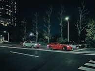 A city street at night with three parked cars under streetlights. Two sedan cars and an SUV are parked side by side, with surrounding trees and tall buildings in the background.