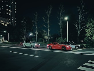 A city street at night with three parked cars under streetlights. Two sedan cars and an SUV are parked side by side, with surrounding trees and tall buildings in the background.