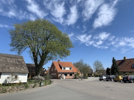A quaint village street features traditional houses with thatched and red-tiled roofs. A large, leafy tree stands prominently in the foreground, surrounded by well-maintained gardens. Several cars are parked along the street, while the sky is blue with scattered white clouds.