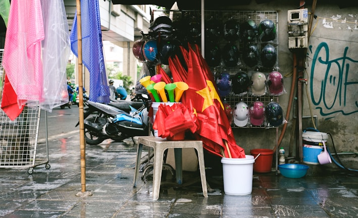 A street-side stall displays multiple red flags with a yellow star, along with colorful plastic horns. A variety of helmets are hung on a wall, and there are plastic buckets and bowls on the ground. Nearby, a scooter is parked beside hanging plastic raincoats in pink and blue. The scene also features a bit of graffiti on the wall.