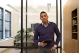 A person stands in a modern office setting holding a tablet. The office has glass walls and large windows that allow natural light to brighten the space. There are plants and a conference table visible in the background, and a wooden shelf is seen to the right.