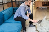 A person is interacting with a laptop while seated on a vibrant blue couch in a modern office setting. The individual is casually dressed, wearing a two-toned blue shirt and black pants. The office features a light wooden floor and a glass coffee table.