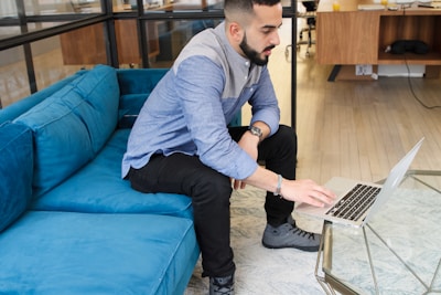 A person is interacting with a laptop while seated on a vibrant blue couch in a modern office setting. The individual is casually dressed, wearing a two-toned blue shirt and black pants. The office features a light wooden floor and a glass coffee table.