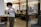 A person with curly hair is taking a photo with their smartphone in a modern office environment. The office features open workspaces with desks, computers, and people working in the background. There are large windows, providing natural light, and the interior includes red piping and minimalist decor.