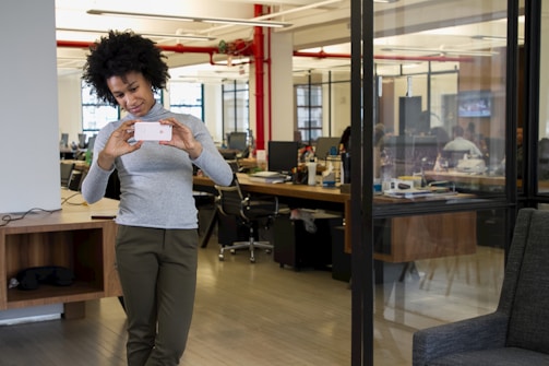 A person with curly hair is taking a photo with their smartphone in a modern office environment. The office features open workspaces with desks, computers, and people working in the background. There are large windows, providing natural light, and the interior includes red piping and minimalist decor.