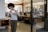 A person with curly hair is taking a photo with their smartphone in a modern office environment. The office features open workspaces with desks, computers, and people working in the background. There are large windows, providing natural light, and the interior includes red piping and minimalist decor.