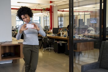 A person with curly hair is taking a photo with their smartphone in a modern office environment. The office features open workspaces with desks, computers, and people working in the background. There are large windows, providing natural light, and the interior includes red piping and minimalist decor.