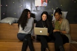 Three women are sitting on wooden benches in a cozy, informal setting. They are gathered around a laptop, seemingly engaged in a collaborative discussion or presentation. They are smiling and appear to be in a relaxed and friendly environment. Behind them is a dark wall with colorful dots and some posters pinned on it. Cushions are placed on the benches, adding to the comfort of the space.