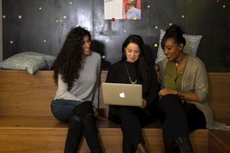 Three women are sitting on wooden benches in a cozy, informal setting. They are gathered around a laptop, seemingly engaged in a collaborative discussion or presentation. They are smiling and appear to be in a relaxed and friendly environment. Behind them is a dark wall with colorful dots and some posters pinned on it. Cushions are placed on the benches, adding to the comfort of the space.
