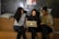 Three women are sitting on wooden benches in a cozy, informal setting. They are gathered around a laptop, seemingly engaged in a collaborative discussion or presentation. They are smiling and appear to be in a relaxed and friendly environment. Behind them is a dark wall with colorful dots and some posters pinned on it. Cushions are placed on the benches, adding to the comfort of the space.