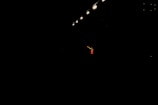 A single performer stands in dramatic lighting, surrounded by darkness. Overhead, several bright stage lights cast a focused beam onto the person, emphasizing their presence. The performer is wearing a red outfit and gesturing with an outstretched arm.