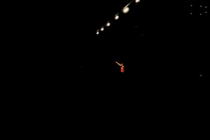 A single performer stands in dramatic lighting, surrounded by darkness. Overhead, several bright stage lights cast a focused beam onto the person, emphasizing their presence. The performer is wearing a red outfit and gesturing with an outstretched arm.
