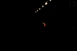 A single performer stands in dramatic lighting, surrounded by darkness. Overhead, several bright stage lights cast a focused beam onto the person, emphasizing their presence. The performer is wearing a red outfit and gesturing with an outstretched arm.