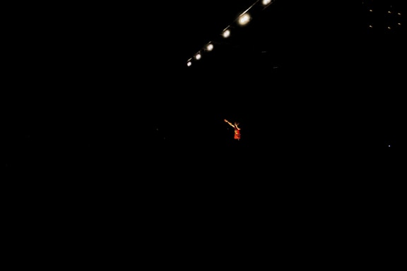 A single performer stands in dramatic lighting, surrounded by darkness. Overhead, several bright stage lights cast a focused beam onto the person, emphasizing their presence. The performer is wearing a red outfit and gesturing with an outstretched arm.