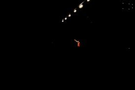 A single performer stands in dramatic lighting, surrounded by darkness. Overhead, several bright stage lights cast a focused beam onto the person, emphasizing their presence. The performer is wearing a red outfit and gesturing with an outstretched arm.