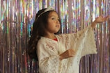 A young child dressed in a white lace outfit with a decorative tiara on their head is posed gracefully against a shimmering backdrop of iridescent streamers. The child has long, dark hair and appears to be gesturing softly with one hand raised.