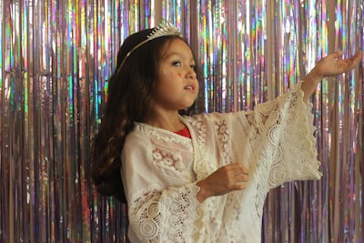 A young child dressed in a white lace outfit with a decorative tiara on their head is posed gracefully against a shimmering backdrop of iridescent streamers. The child has long, dark hair and appears to be gesturing softly with one hand raised.