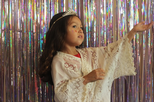 A young child dressed in a white lace outfit with a decorative tiara on their head is posed gracefully against a shimmering backdrop of iridescent streamers. The child has long, dark hair and appears to be gesturing softly with one hand raised.