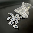 A collection of small hamsa hand charms with a central blue eye design spread out on a sleek black surface. Several charms are spilling out from an open plastic pouch. The charms are metallic and intricately detailed.