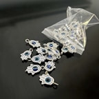 A collection of small hamsa hand charms with a central blue eye design spread out on a sleek black surface. Several charms are spilling out from an open plastic pouch. The charms are metallic and intricately detailed.