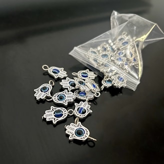 A collection of small hamsa hand charms with a central blue eye design spread out on a sleek black surface. Several charms are spilling out from an open plastic pouch. The charms are metallic and intricately detailed.