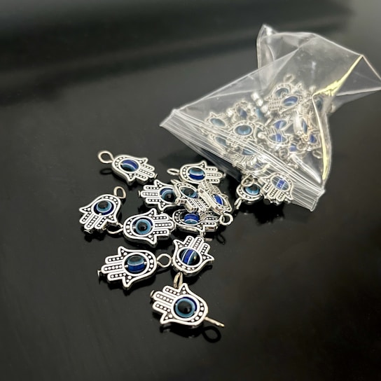 A collection of small hamsa hand charms with a central blue eye design spread out on a sleek black surface. Several charms are spilling out from an open plastic pouch. The charms are metallic and intricately detailed.