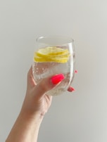 A hand with bright pink nail polish holds a clear glass containing sparkling water with lemon slices. The background is a plain light gray, creating a minimalistic and refreshing appearance.