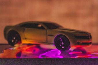 A toy car positioned on a surface with colorful lights creating a vibrant reflection. The vehicle appears to be a miniature model of a sports car, highlighted with purple and orange lighting effects. The background is blurred and neutral, focusing attention on the car.
