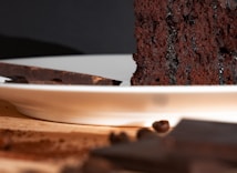 A close-up of a rich, moist chocolate cake slice on a white plate, with a piece of dark chocolate and coffee beans in the foreground, set against a dark background.