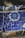 A close-up view of a mosaic wall composed of various ceramic tiles. The central feature is a circular decorative tile with intricate blue patterns. Surrounding it are irregularly shaped pieces of blue and beige tiles, set in grout.