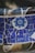 A close-up view of a mosaic wall composed of various ceramic tiles. The central feature is a circular decorative tile with intricate blue patterns. Surrounding it are irregularly shaped pieces of blue and beige tiles, set in grout.