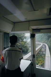 A person in uniform is standing inside the driver's cabin of a train, looking out over a railway track that curves into the distance through a lush green landscape.