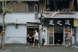 An urban street scene with a small business displaying signage in a foreign script. People are casually walking past a shop with an open exterior and visible interior, where customers can be seen inside. The facade includes plants, an air conditioning unit, and worn architecture, suggesting an aged building. A tree stands prominently in the foreground, partially wrapped with material.