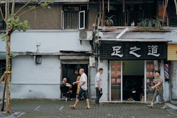 An urban street scene with a small business displaying signage in a foreign script. People are casually walking past a shop with an open exterior and visible interior, where customers can be seen inside. The facade includes plants, an air conditioning unit, and worn architecture, suggesting an aged building. A tree stands prominently in the foreground, partially wrapped with material.