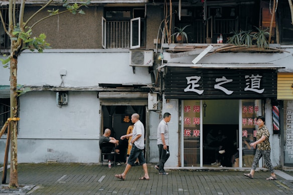 An urban street scene with a small business displaying signage in a foreign script. People are casually walking past a shop with an open exterior and visible interior, where customers can be seen inside. The facade includes plants, an air conditioning unit, and worn architecture, suggesting an aged building. A tree stands prominently in the foreground, partially wrapped with material.