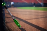 A green and black baseball bat is leaning against a fence on a baseball field. The field has a dirt section in the foreground and green grass in the background. The seating area with red seats is visible in the background, and a couple of blurred figures can be seen on the field.