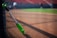 A green and black baseball bat is leaning against a fence on a baseball field. The field has a dirt section in the foreground and green grass in the background. The seating area with red seats is visible in the background, and a couple of blurred figures can be seen on the field.