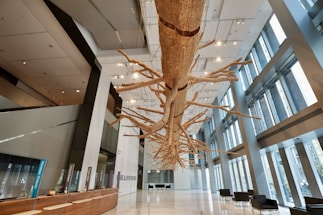 A large, intricately detailed suspended sculpture resembling a tree with numerous branches extends through a modern and spacious lobby with high ceilings and floor-to-ceiling windows. The lobby features a wooden reception desk on the left, illuminated by numerous ceiling lights, and seating areas with black chairs on the right.