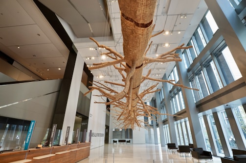 A large, intricately detailed suspended sculpture resembling a tree with numerous branches extends through a modern and spacious lobby with high ceilings and floor-to-ceiling windows. The lobby features a wooden reception desk on the left, illuminated by numerous ceiling lights, and seating areas with black chairs on the right.