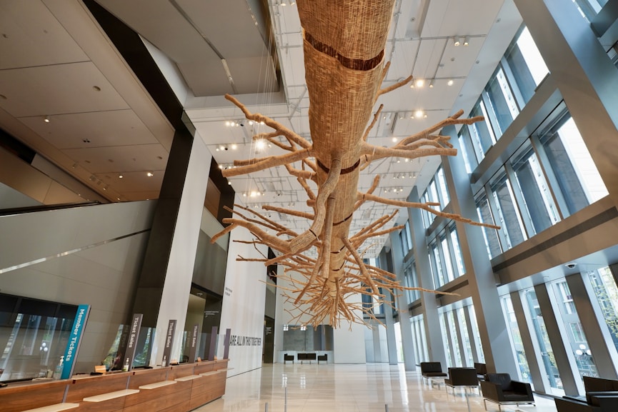 A large, intricately detailed suspended sculpture resembling a tree with numerous branches extends through a modern and spacious lobby with high ceilings and floor-to-ceiling windows. The lobby features a wooden reception desk on the left, illuminated by numerous ceiling lights, and seating areas with black chairs on the right.