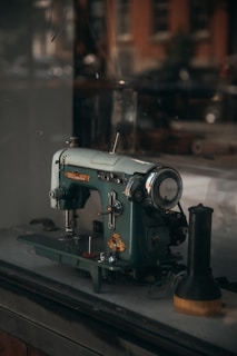 A vintage sewing machine is sitting on a table, showcasing intricate metal details and a robust, mechanical design. The lighting is soft and warm, creating a nostalgic atmosphere, while the blurred background suggests an old shop or studio setting.