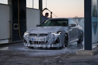 A person is washing a silver BMW car with high-pressure water at a car wash station. The car is covered in soap suds, and the sun appears to be setting or rising in the background.